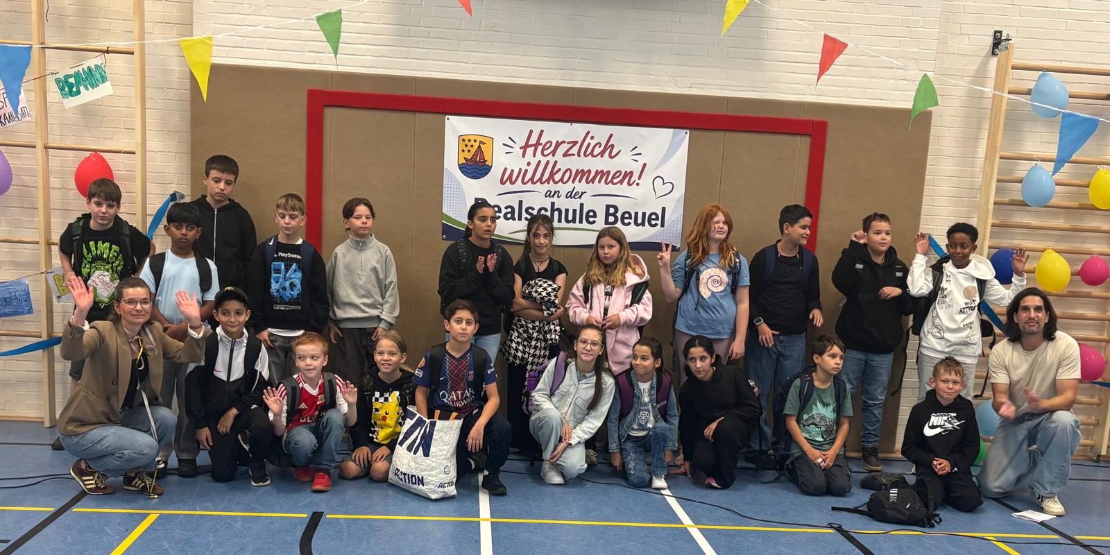 Eine neue 5. Klasse mit ihrem Klassenlehrerteam vor dem Banner „Herzlich willkommen an der Realschule Beuel“.