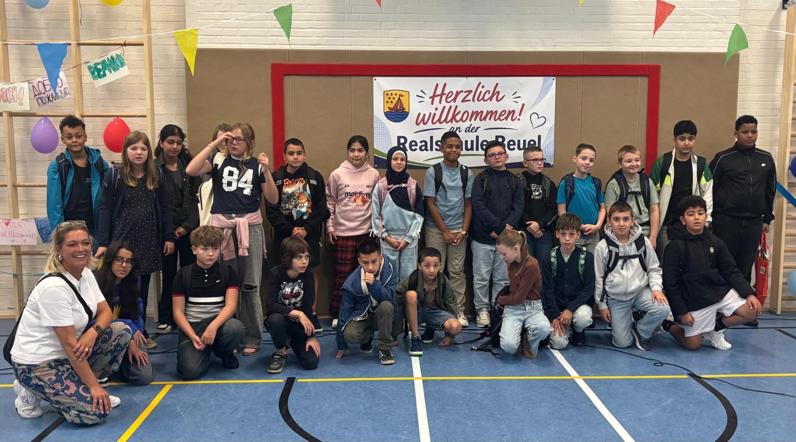 Eine neue 5. Klasse mit Klassenlehrerin vor dem Willkommensbanner in der Sporthalle.