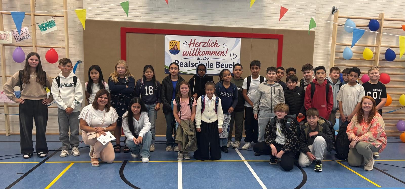 Eine neue 5. Klasse mit ihrem Klassenlehrerteam vor dem Willkommensbanner in der Sporthalle.