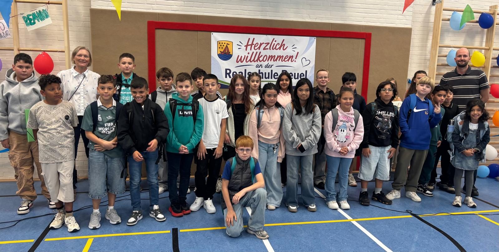 Eine neue 5. Klasse mit ihrem Klassenlehrerteam vor dem Willkommensbanner in der Sporthalle.